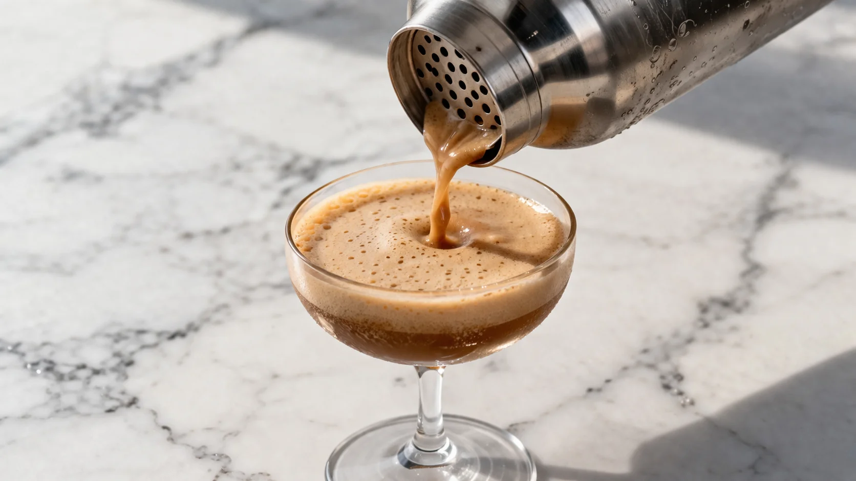 Shaker en inox versant un café shakerato protéiné mousseux dans un verre à cocktail transparent, mousse brune visible, fond de marbre blanc