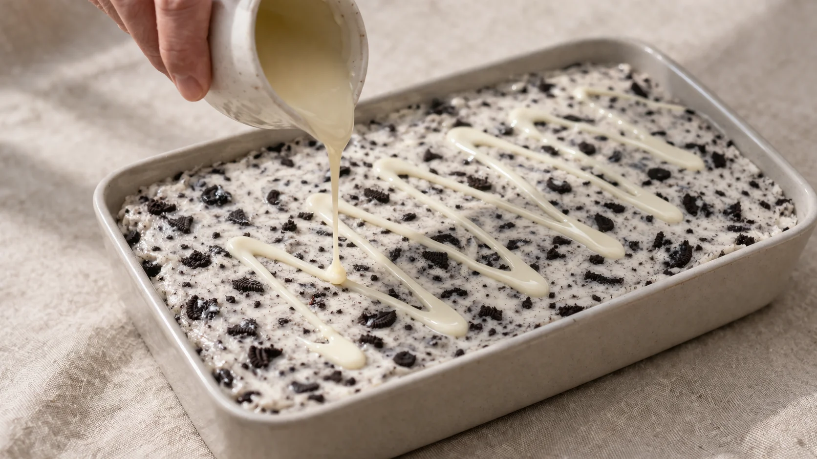 Fond de verre comprimant la masse cookies and cream blanche mouchetée de noir dans un plat rectangulaire tapissé de papier cuisson, éclats de biscuits posés sur la surface