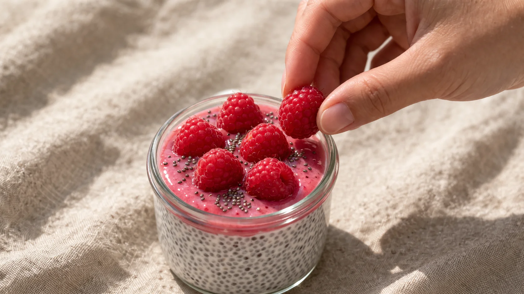 Main disposant des framboises fraîches sur le dessus d'un chia pudding dans un bocal en verre, coulis rose vif et graines de chia saupoudrées visibles, fond lin beige