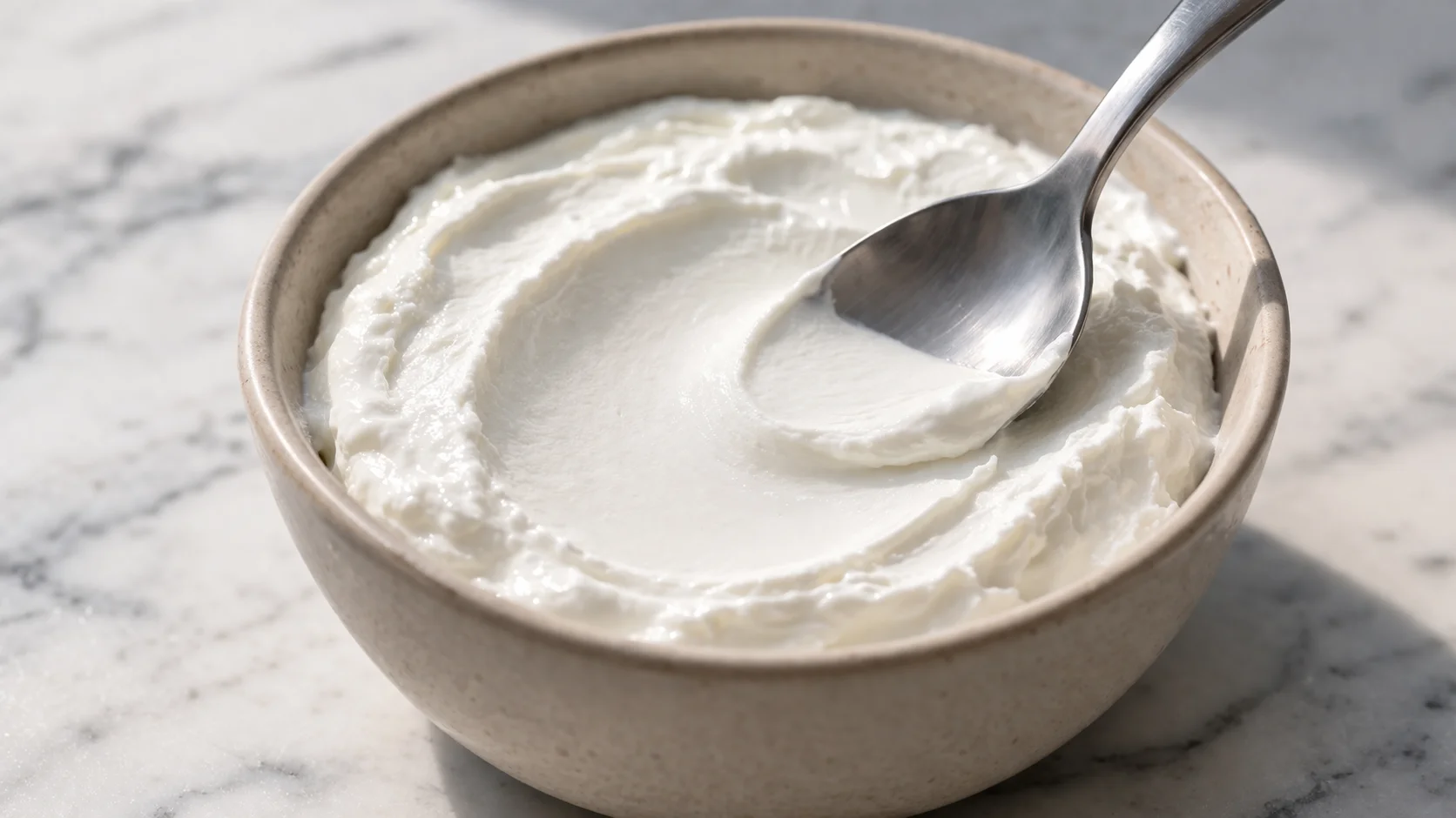 Cuillère lissant le yaourt grec épais et blanc dans un bol en céramique beige, texture crémeuse et épaisse bien visible, plan de travail en marbre