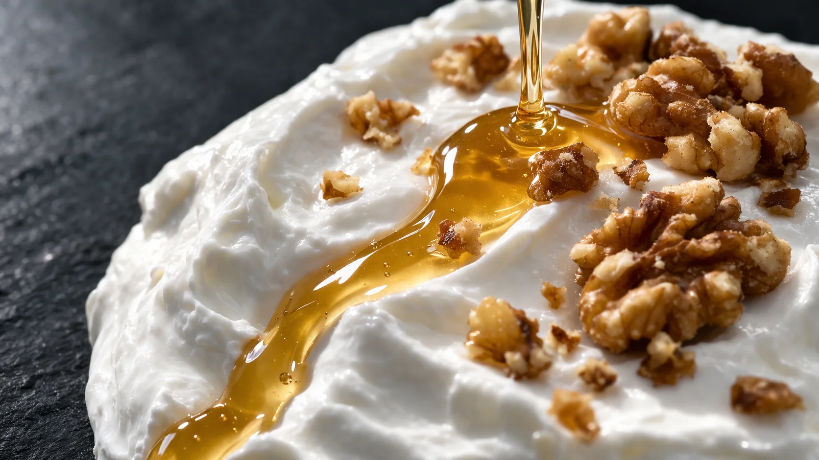 Gros plan sur la texture épaisse et crémeuse d'un yaourt grec avec un filet de miel doré brillant et des morceaux de noix croquants, fond ardoise sombre