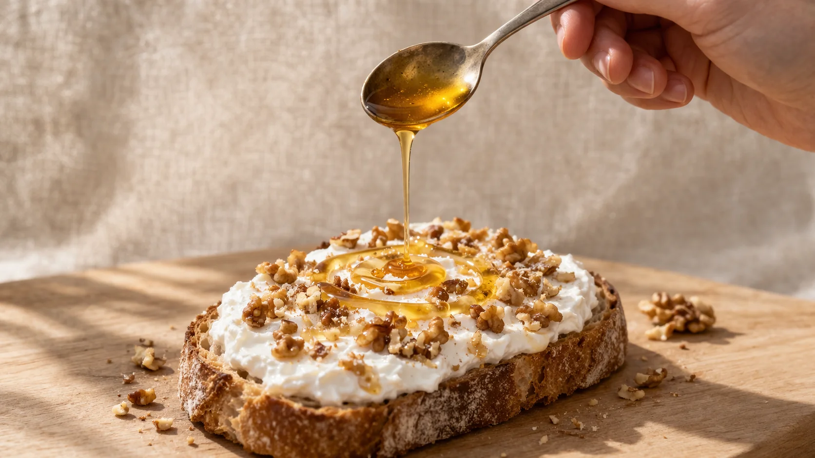 Main versant un filet de miel doré sur une tartine de pain complet généreusement garnie de fromage blanc épais et de cerneaux de noix concassés, fond lin beige