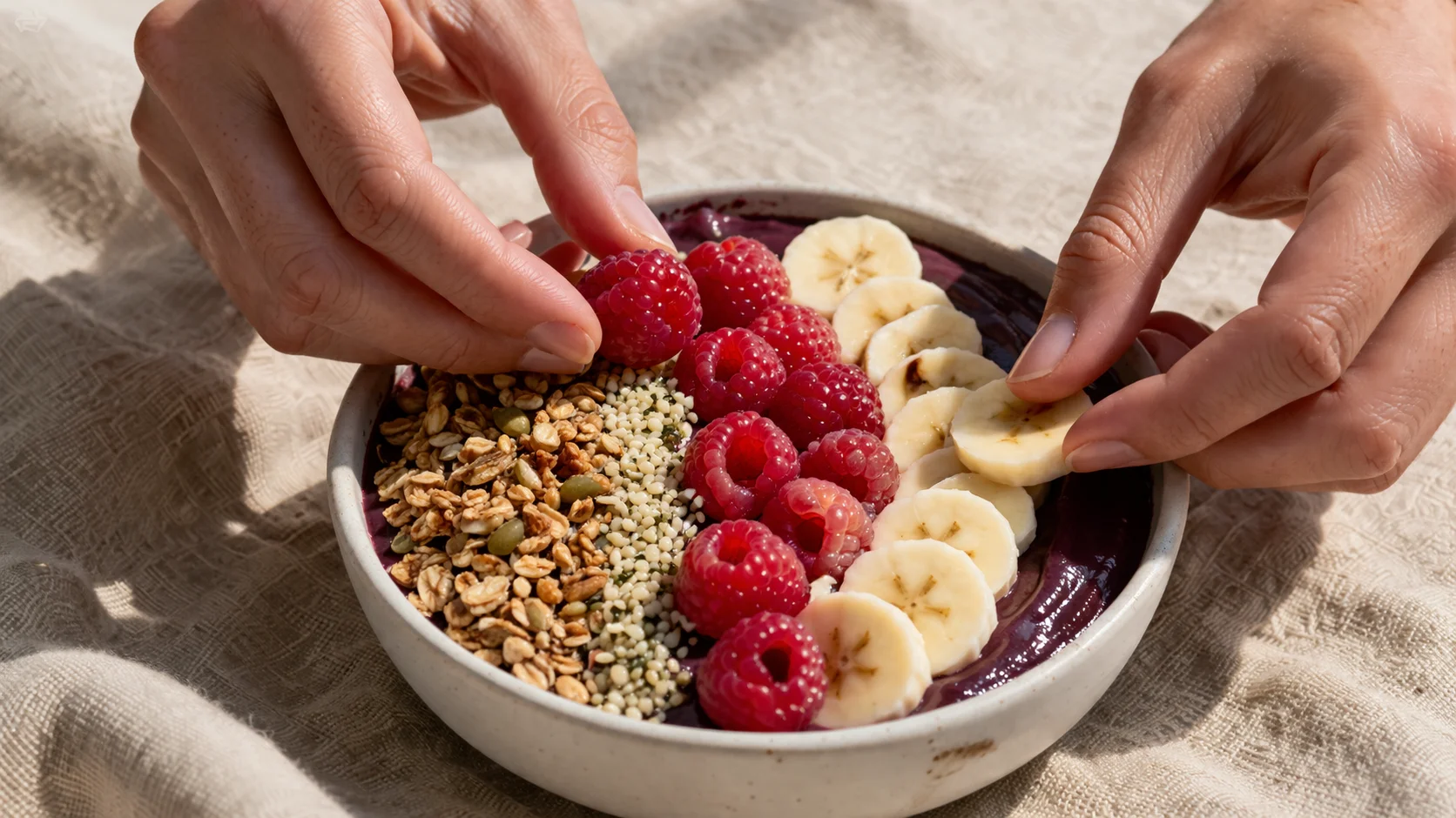 Mains disposant des framboises fraîches et des rondelles de banane sur la surface violette épaisse d'un bowl açaï dans un bol en céramique blanche, granola et graines visibles