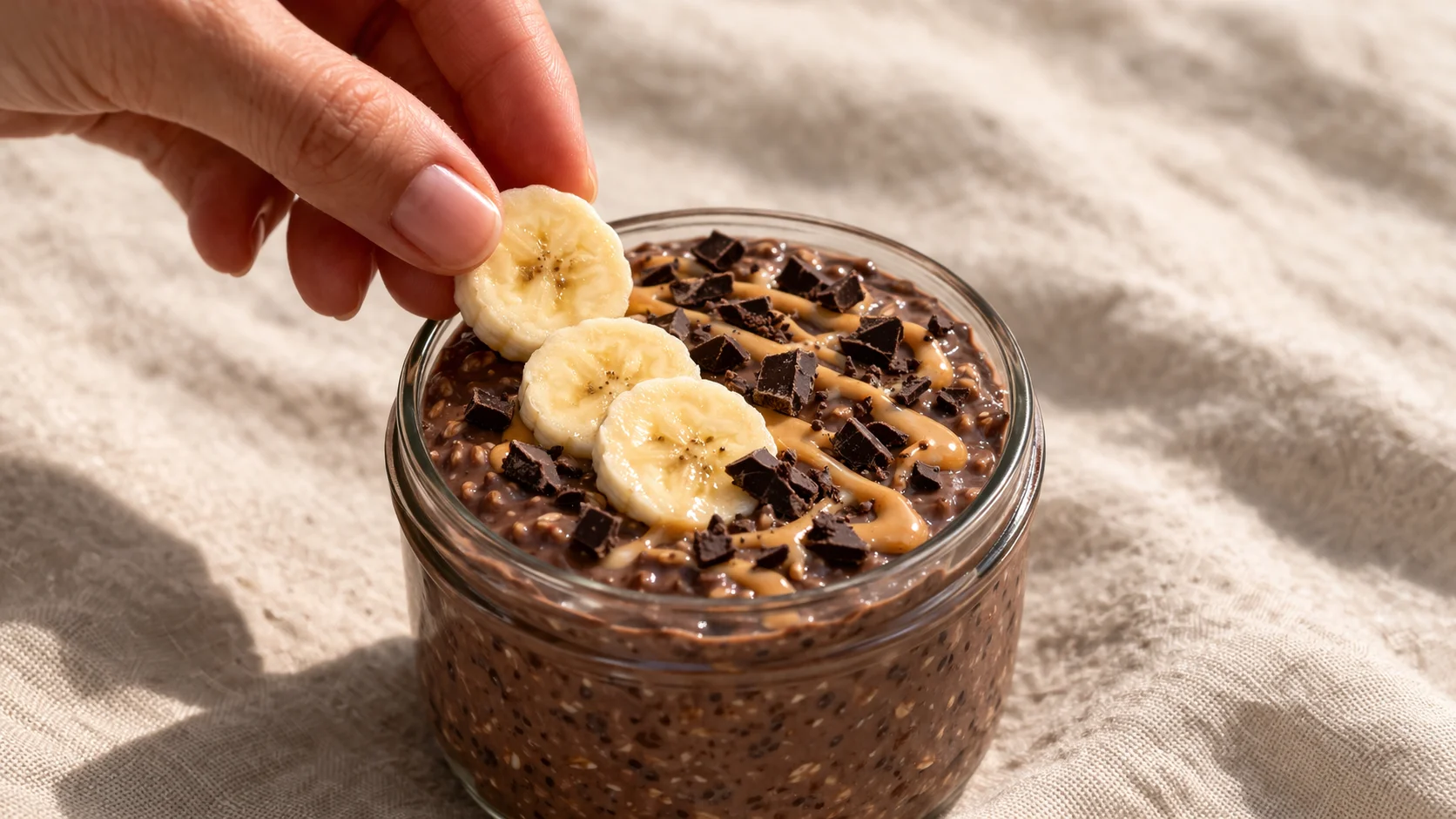 Main disposant des rondelles de banane fraîche sur des overnight oats chocolat crémeux dans un bocal en verre, éclats de chocolat noir et filet de beurre de cacahuète visibles, fond lin beige
