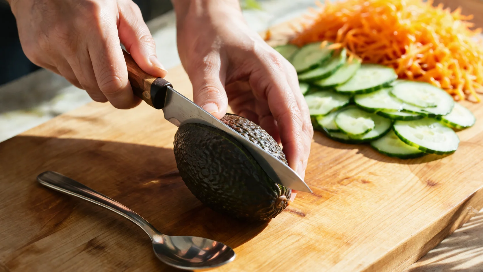 Mains tranchant un avocat mûr en deux sur une planche, cuillère prête à le détacher de la peau, autres garnitures colorées disposées autour