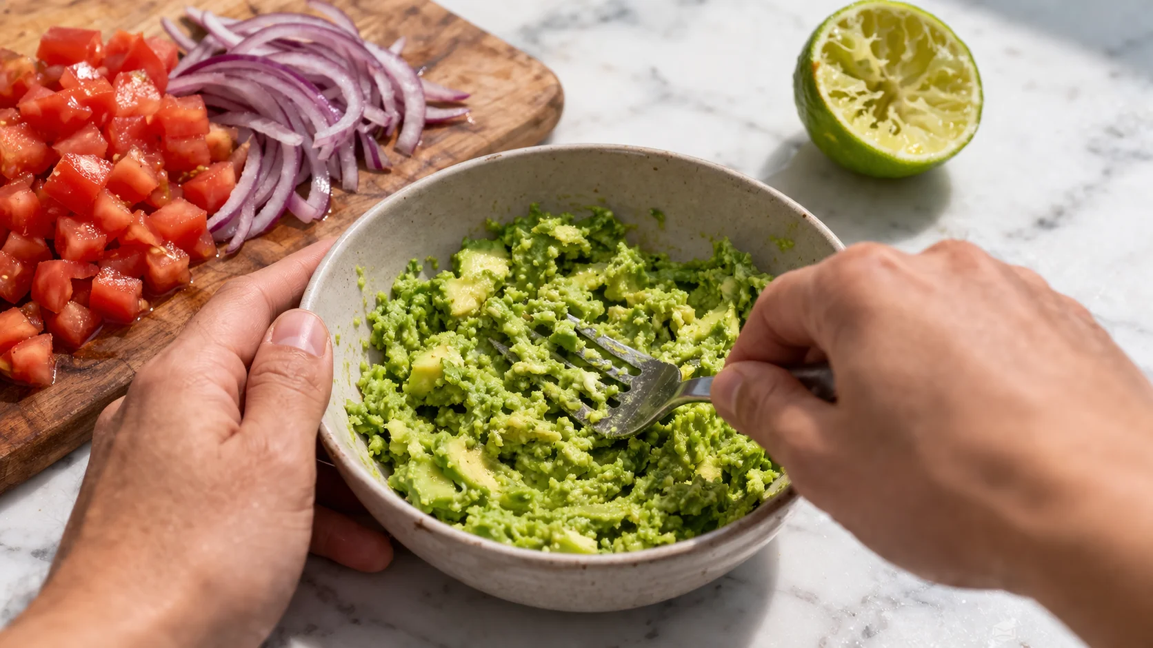 Fourchette en train d'écraser grossièrement la chair d'avocat dans un bol, dés de tomate et oignon rouge visibles, citron vert à côté